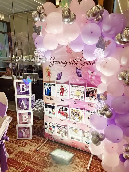 A "Growing with Grace" milestone photo board for a first birthday. The display is framed by a beautiful garland of purple and pink balloons, with "BABY" blocks adding a classic touch.