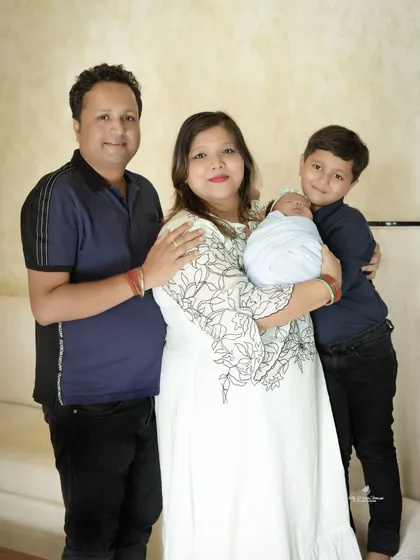 A full-length portrait of a family of four. The parents and older son stand together, smiling warmly, as they introduce their newborn baby.