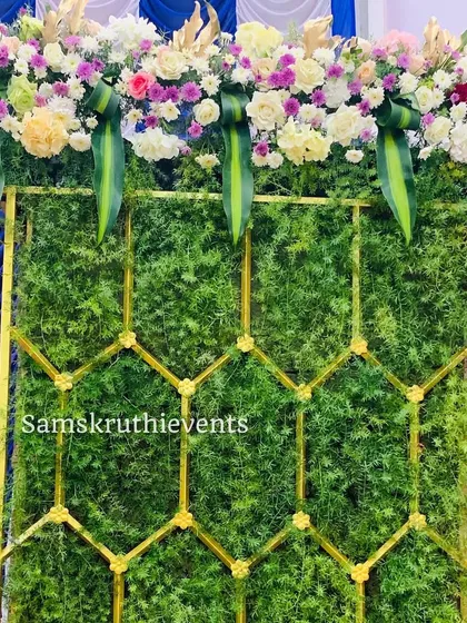 A modern backdrop panel made of a lush green wall and a gold geometric frame. The top is decorated with a beautiful arrangement of pastel-colored flowers.