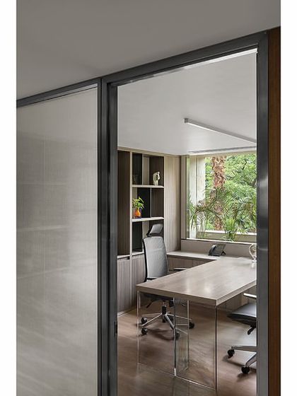 A view into a cabin from the main corridor in 'The Layered Office'. The design emphasizes transparency and connection, with glass walls allowing natural light to flow from the exterior-facing cabins into the central circulation areas.