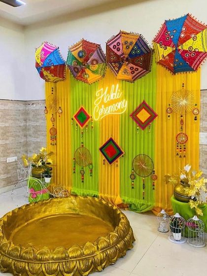 A trendy Haldi ceremony setup with a 'Haldi Ceremony' neon sign. The backdrop uses lime green and yellow drapes, decorated with colorful Rajasthani umbrellas and traditional hangings for a fusion look.