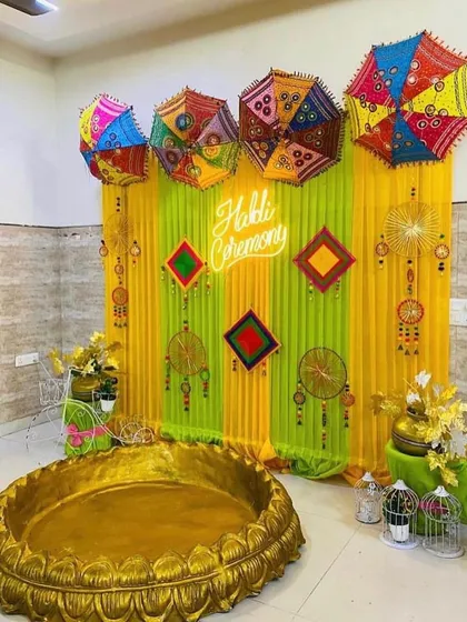 A trendy Haldi ceremony setup with a 'Haldi Ceremony' neon sign. The backdrop uses lime green and yellow drapes, decorated with colorful Rajasthani umbrellas and traditional hangings for a fusion look.