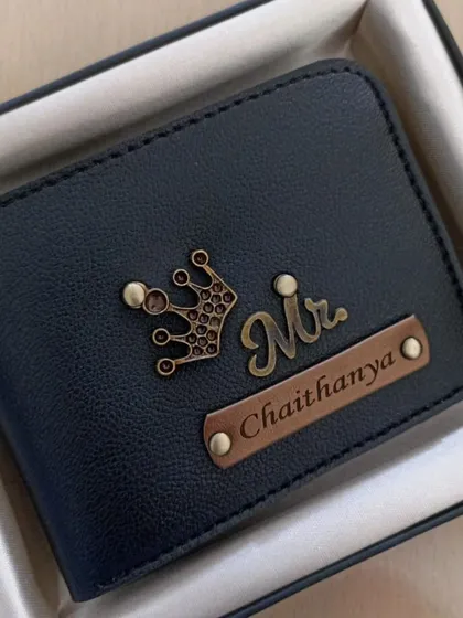 A close-up of a personalized navy blue wallet with a 'Mr.' and crown charm, showcasing the fine details of the engraving and charms.