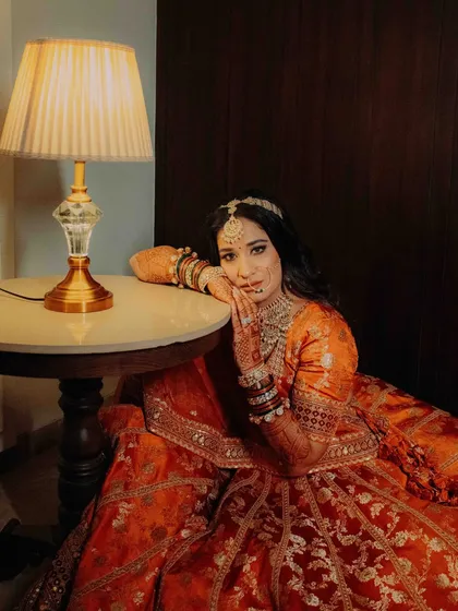 A quiet, posed portrait of the bride. She rests by a table, looking at the camera, showcasing her intricate lehenga and jewelry in a moment of calm.