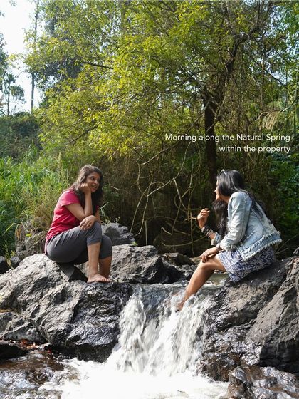 Two women chat while dipping their feet in a natural spring in Coorg. My trips include stays at properties with such private, serene spots for you to unwind.