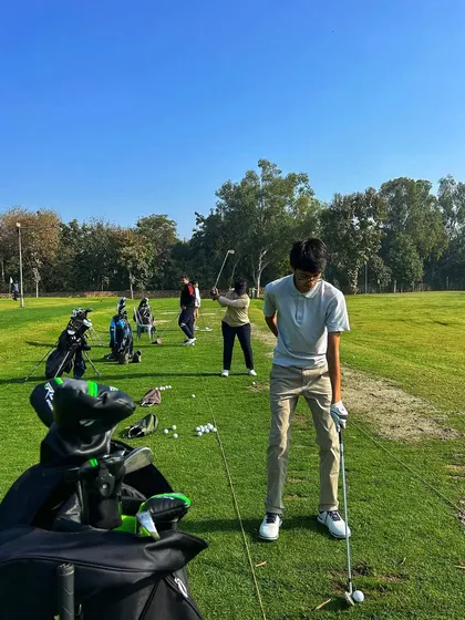 A young golfer in perfect setup position, with his golf bag in the foreground and other players on the range behind him.