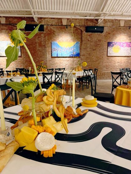 A wide view of our edible art tablescape. The centerpiece of bread, cheese, and sunflowers sits on a custom-printed graphic tablecloth, creating a striking contrast with the rustic brick walls of the art gallery venue.