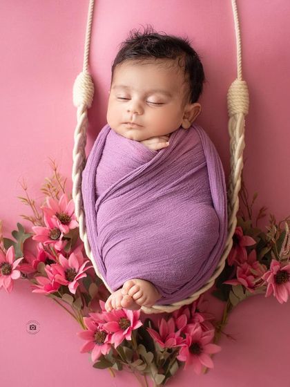 A newborn rests in a hanging swing, wrapped in a lovely lavender swaddle and surrounded by pink flowers.