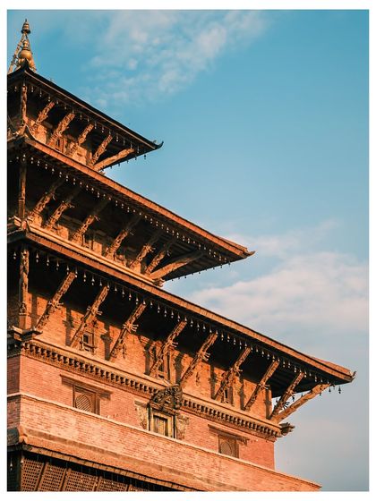 The detailed, multi-tiered roof of a traditional Newari pagoda in Kathmandu. The warm sunlight highlights the intricate wooden carvings against a brilliant blue sky.
