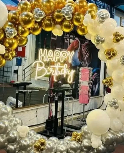A birthday celebration in a gym. This decoration features a circular balloon arch in gold, silver, and white, with a "Happy Birthday" neon sign.