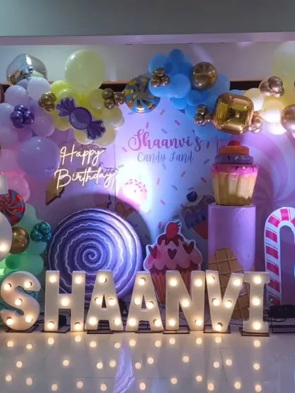 Shaanvi's Candyland party setup with large marquee letters, a custom backdrop, and props like giant cupcakes and a swirl lollipop, all illuminated for a magical effect.