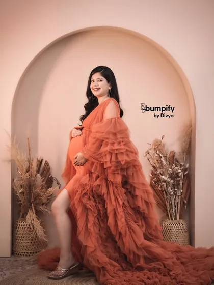 A vibrant and joyful portrait of a mom-to-be in a fiery orange ruffled gown. The simple arched backdrop and boho props complement the dress beautifully.