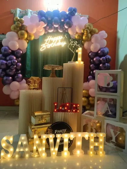 A sophisticated birthday setup with a balloon ring in shades of purple and gold, a 'Happy Birthday' neon sign, and the name 'Sanvith' in marquee letters.