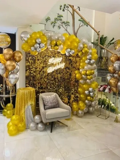 A luxurious gold and silver birthday setup. A shimmering gold sequin wall is framed by a matching balloon arch, creating a glamorous photo booth area in a home.