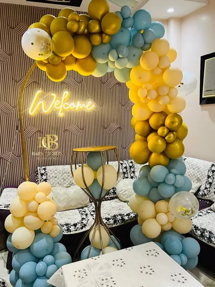 A cheerful 'Welcome' setup for a new baby boy. The decor features a balloon arch in shades of yellow and blue, a neon sign, and an elegant pedestal for placing gifts or a cake.