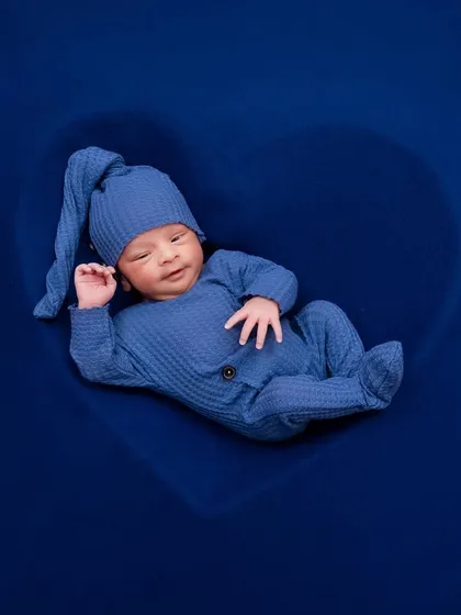 A sweet, sleepy smile from a newborn boy in a blue outfit, nestled inside a heart prop.