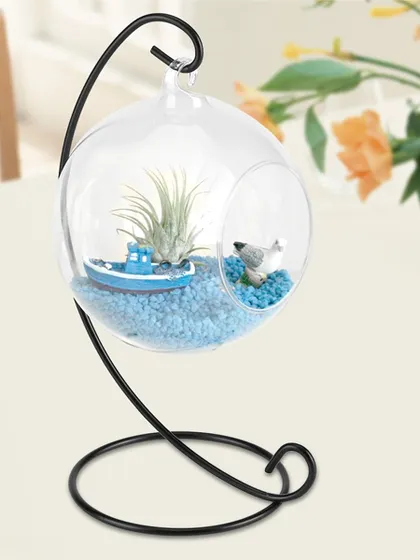 This hanging glass bowl terrarium, complete with an iron stand, is a delightful way to display air plants or small succulents. The setup includes decorative blue sand and miniature boat and bird figures, creating a tiny coastal scene.