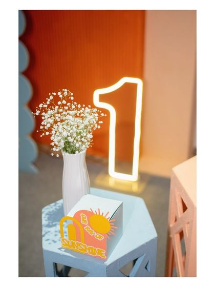 A detail from a "First Trip Around the Sun" party. A modern, light-up number '1' sign is paired with a custom 'You Are My Sunshine' box and a vase of baby's breath.