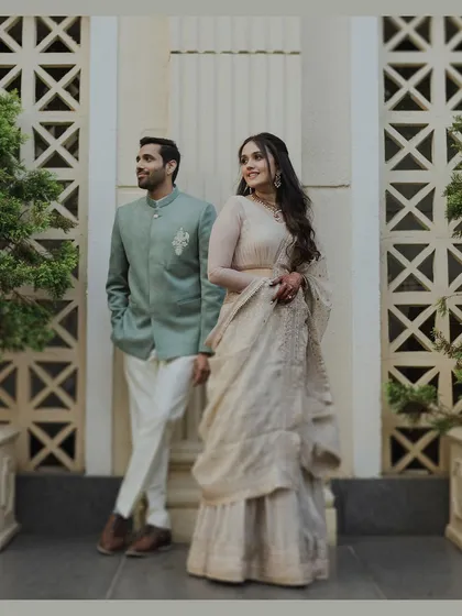 A full-length portrait of an elegantly dressed couple. The groom in a mint green bandhgala and the bride in a cream lehenga stand against a modern, patterned wall, showcasing a timeless and stylish engagement look.