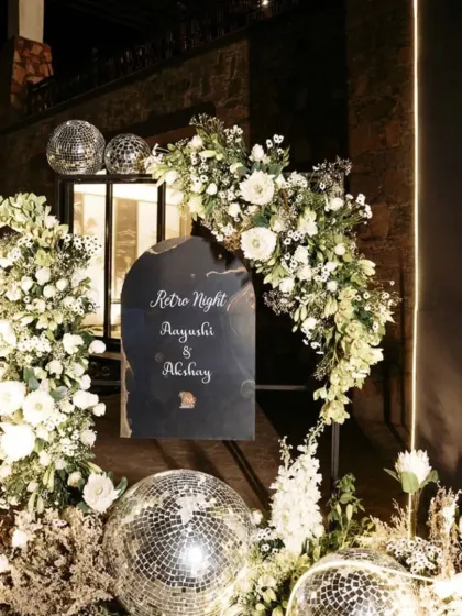 A "Retro Night" themed entrance for a Sangeet party. The decor includes a custom welcome sign, disco balls, and an arch of white flowers, setting a fun and nostalgic tone for the evening.