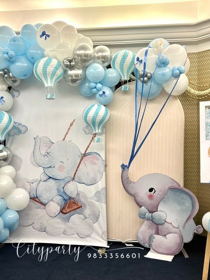 A beautiful naming ceremony decoration with a baby elephant and hot air balloon theme. The setup includes a custom backdrop, a blue, white, and silver balloon arch, and an easel for the name reveal.