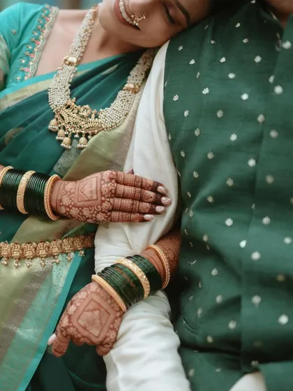 A detailed shot focusing on the bride's hands, adorned with intricate henna and traditional green bangles, resting on the groom's arm. It highlights the fine details of the saree border, kamarbandh, and the texture of the groom's jacket.