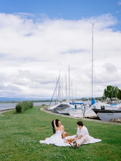 A perfect picnic scene by the water in Switzerland. This wide shot captures the serenity and beauty of their chosen spot, with the sailboats adding to the idyllic atmosphere.
