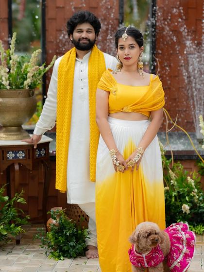 A modern couple's look for a pre-wedding event. Her yellow and white draped outfit is perfectly matched with his white kurta and bright yellow scarf.