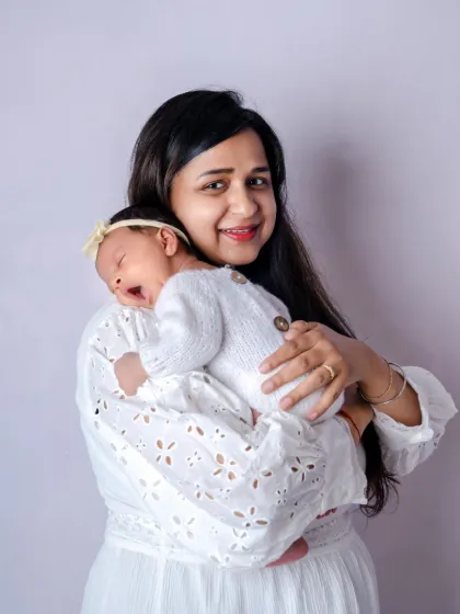 A mother smiles warmly, holding her sleeping newborn close. The soft, all white theme of this studio newborn photo highlights the purity and peace of these early days.