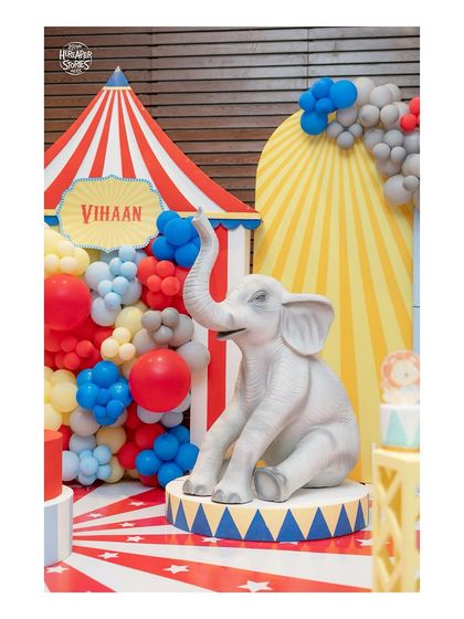 A close-up of a circus-themed setup, featuring a life-sized elephant prop on a pedestal in front of a classic red-and-white striped tent.