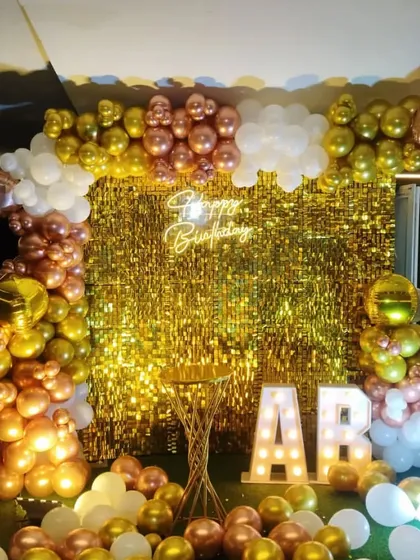 A birthday party setup with a gold shimmer wall, a balloon arch in gold, rose gold, and white, and marquee letters.