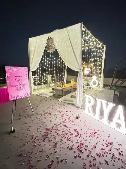 An elegant bachelorette party on a rooftop, complete with a draped canopy, fairy light curtains, personalized marquee letters, and scattered rose petals for a luxurious feel.