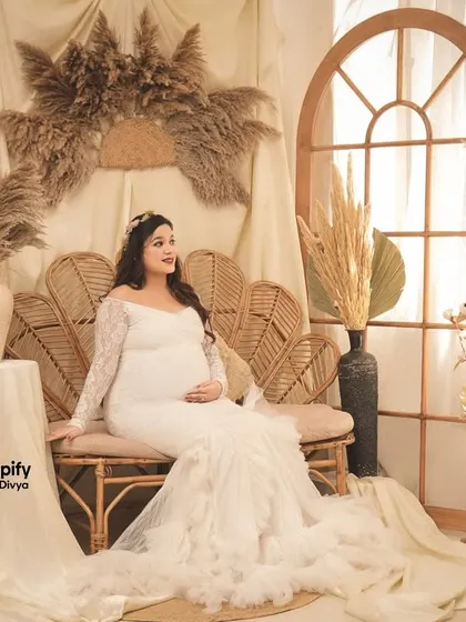 A beautiful, light-filled portrait in our boho corner. The mom-to-be is seated on a wicker sofa in a lovely white lace gown, surrounded by pampas grass and soft fabrics.