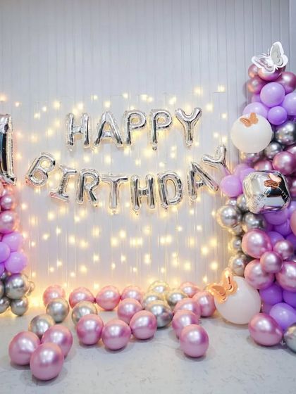 A simple wall decoration with pink and silver balloons, fairy lights, and "Happy Birthday" foil letters.