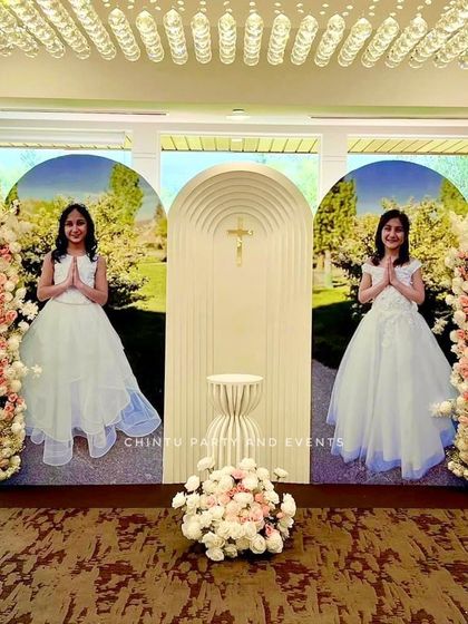 A Holy Communion flower decoration with large photo panels of the children. The setup uses a modern white arch and tall pink and white floral arrangements.