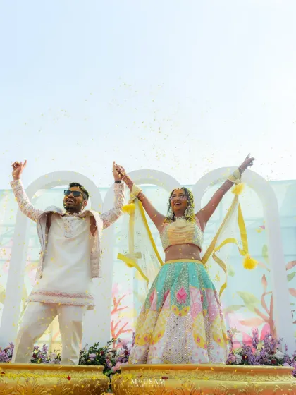 A triumphant moment at a Haldi ceremony, set against a beautiful pastel backdrop. The couple raises their hands in celebration, showered with confetti, perfectly showcasing how great decor can elevate the energy of a photograph.