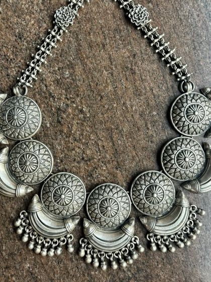 An antique-style oxidised silver necklace. This piece features a series of circular and crescent-shaped motifs with ghungroo drops, creating a substantial and eye-catching choker.
