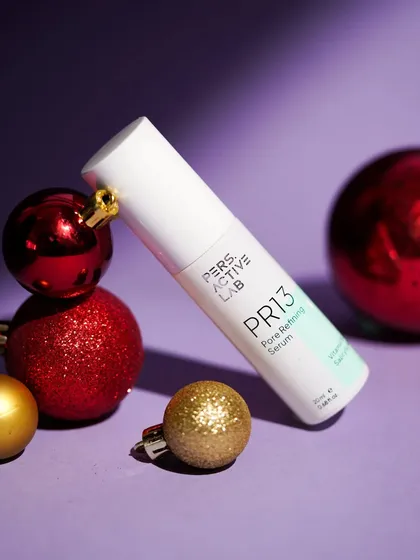 A clean, festive product shot of a pore refining serum placed among shiny red and gold Christmas ornaments. The purple background provides a rich contrast, making the product pop.