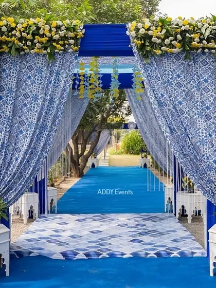 A stunning entrance walkway for a Santorini-themed Haldi. The path is lined with blue carpet and draped in traditional patterned fabric, topped with fresh yellow and white flowers.