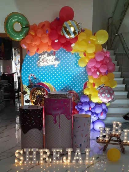 A candy-themed birthday party with a vibrant color palette. The setup includes a polka-dot backdrop, a neon "Happy Birthday" sign, and pedestals designed to look like dripping chocolate and sprinkles.