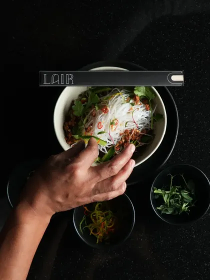 Experience the best blend of pan-Asian cultures. A chef's hand carefully garnishes a bowl of glass noodles, a dish that combines fresh herbs and savory elements.