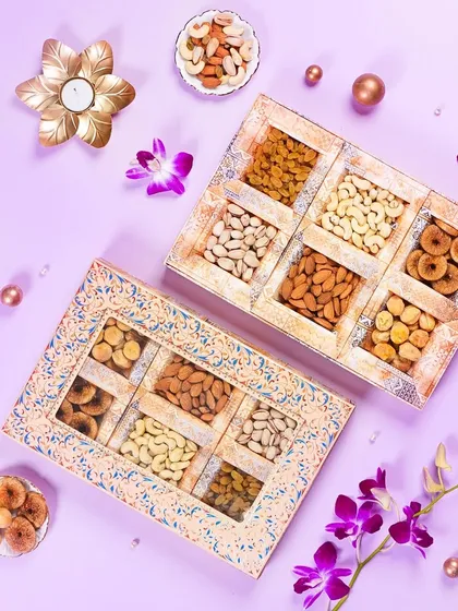 An elegant flatlay of two rustic-style dry fruit gift boxes on a pastel purple background. The scattered flowers and pearls add a delicate and premium touch to the festive packaging.