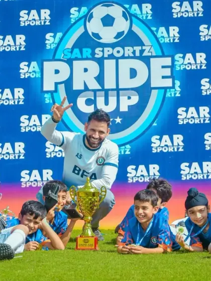 A coach and his team of young players celebrate with their trophy in front of the Sar Sportz Pride Cup banner.