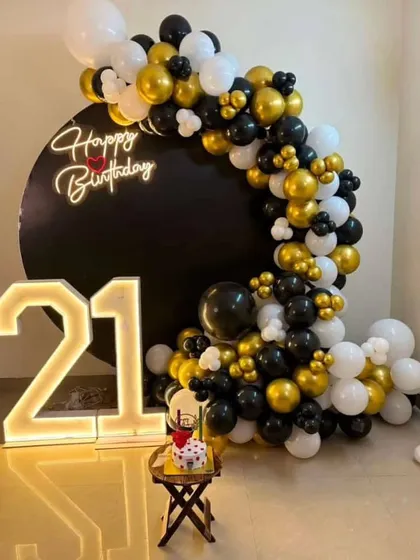 A classic and elegant 21st birthday setup. The black backdrop is accented with a semi-arch of black, gold, and white balloons and a large light-up number '21'.