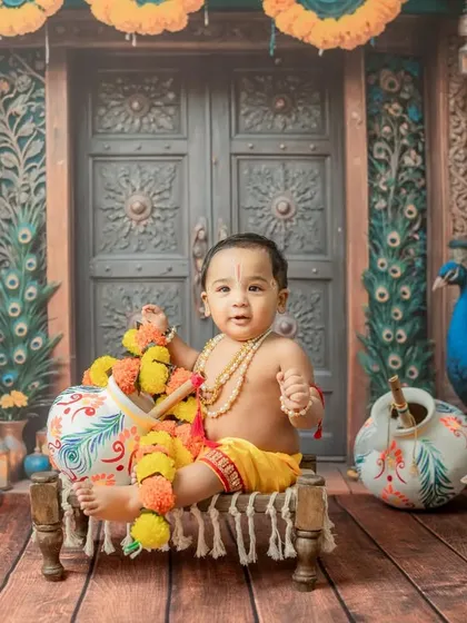 Seated on a small cot, this baby Krishna looks on with a playful expression, with peacock and cow props enhancing the Vrindavan-like scene.