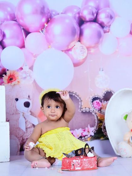 A baby girl sitting with her birthday cake during a pink and yellow themed party setup.