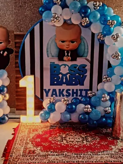 A classic Boss Baby first birthday setup for Yakshit, featuring a character backdrop, marquee number '1', and a blue, white, and silver balloon arch.