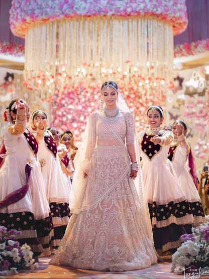 A cinematic shot of the bride's entry, escorted by dancers. Her makeup glows under the grand chandelier and stage lights.