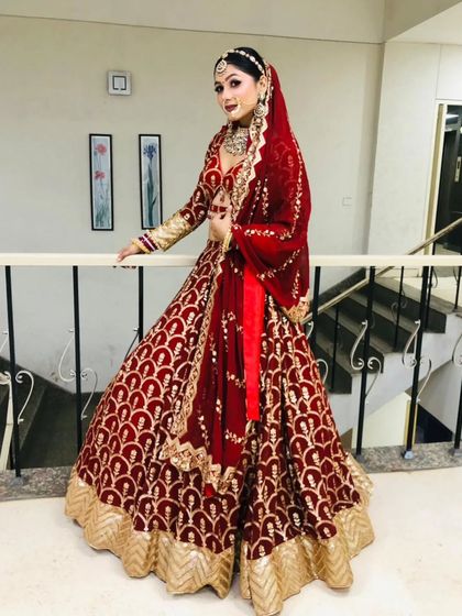 Full-length view of a bride wearing a maroon velvet lehenga with a unique arched pattern, standing by a railing.