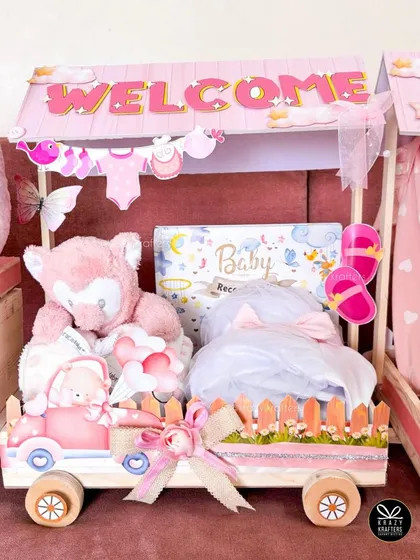 A close-up of one of the carriages from the 'Welcome Baby' train, filled with a soft teddy bear, baby record book, and other adorable pink-themed gifts.